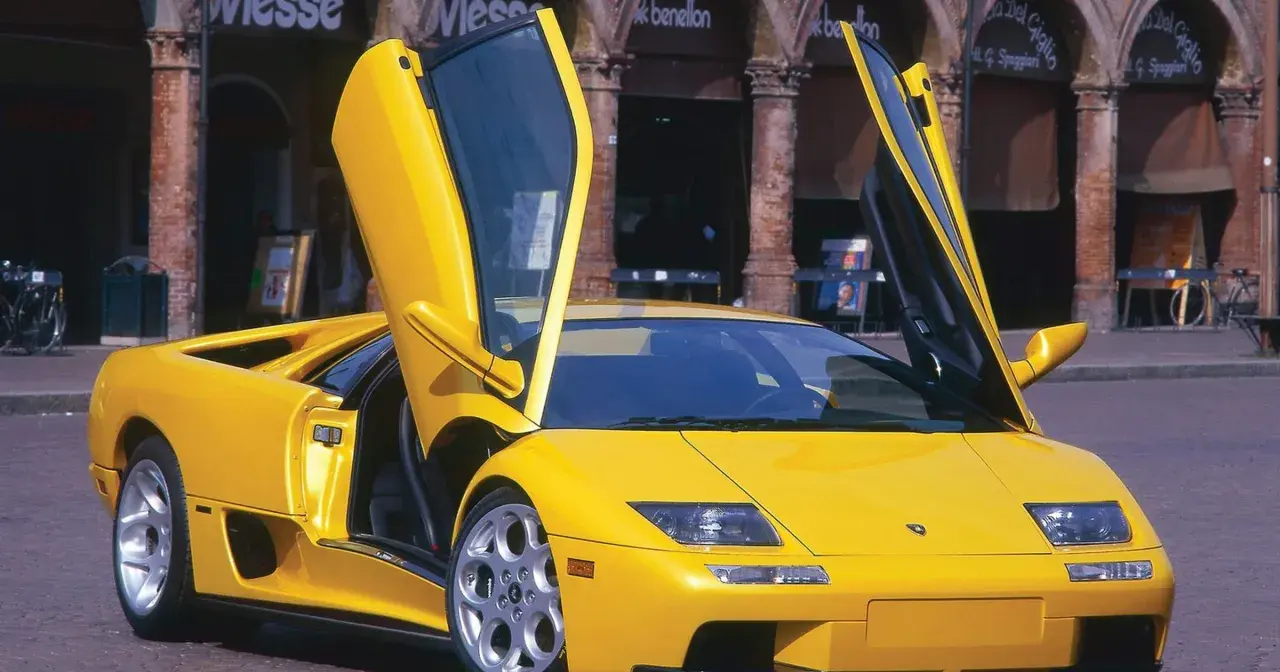 Żółty Lamborghini Diablo z otwartymi drzwiami, zaparkowany na brukowanej ulicy przed budynkami z arkadami.