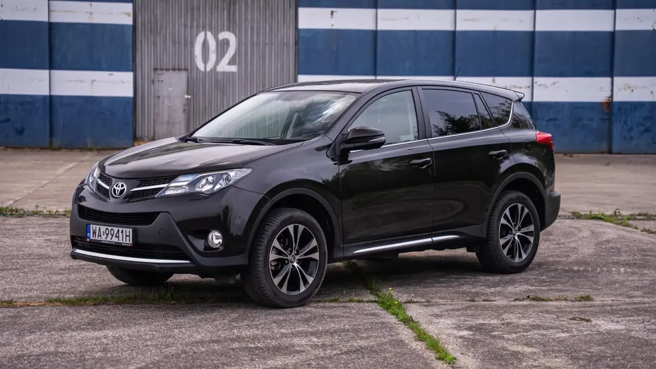 Czarny, używany Toyota RAV4 z salonu, zaparkowany przed budynkiem z niebiesko-białymi pasami.