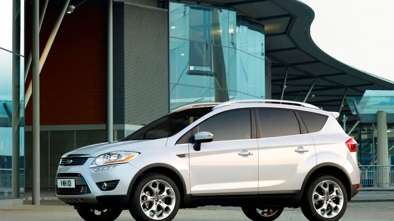 Biały Ford Kuga 2010 zaparkowany przed nowoczesnym budynkiem z przeszkloną fasadą.