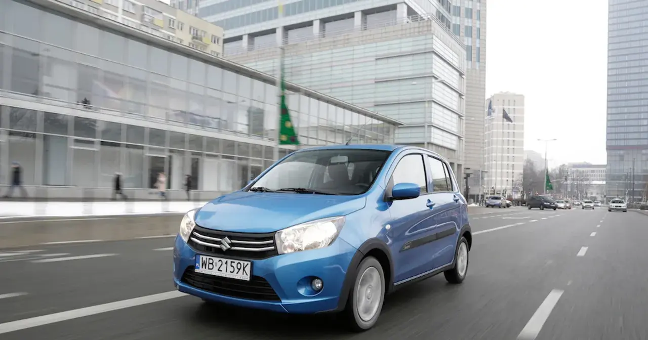 Niebieski Suzuki Celerio jedzie ulicą w mieście, mijając nowoczesne budynki i innych uczestników ruchu.