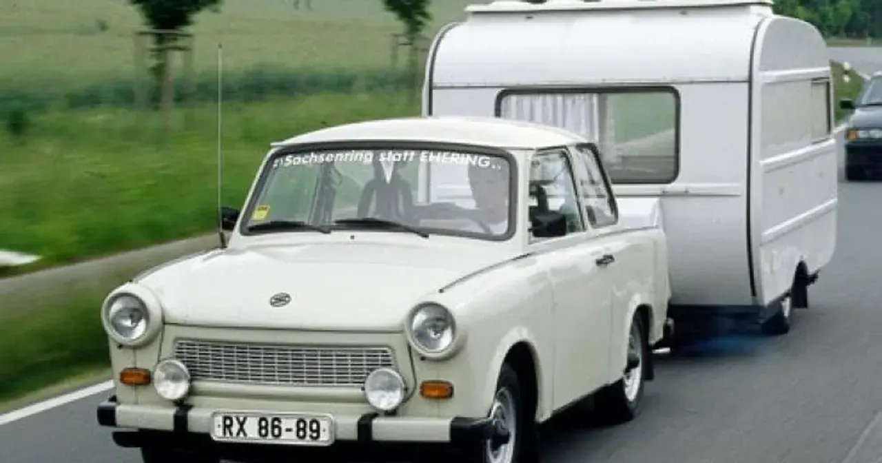 Biały Trabant ciągnie przyczepę kempingową po drodze.