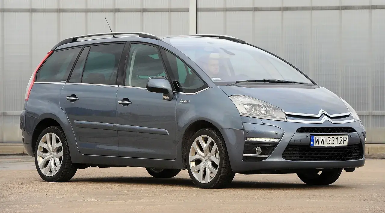 Szary Citroen Picasso z napisem "Picasso" na błotniku, zaparkowany przed budynkiem.