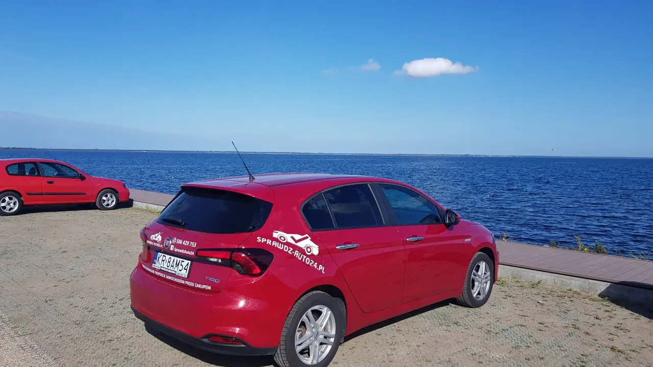 Czerwony fiat tipo sedan z napisem "SprawdzAuto24.pl" stoi nad błękitnym jeziorem w słoneczny dzień.