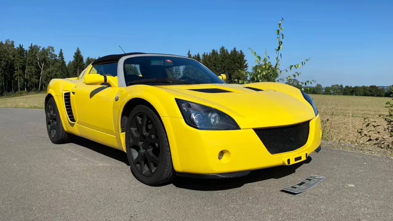 Żółty Opel Speedster na tle lasu i pól. Słońce pięknie podkreśla jego sportową sylwetkę.