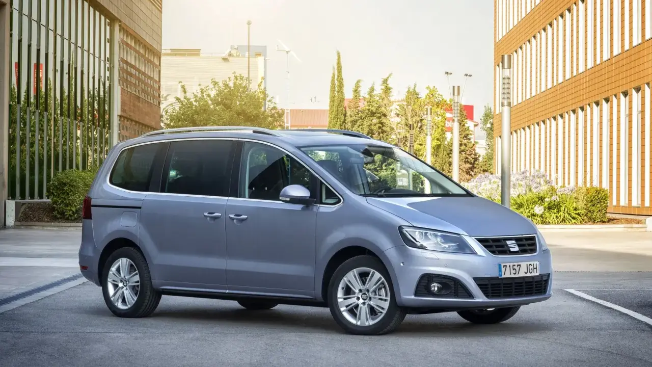 Srebrny Seat Alhambra, rodzinny van, parkuje przed nowoczesnym budynkiem z brązową elewacją.