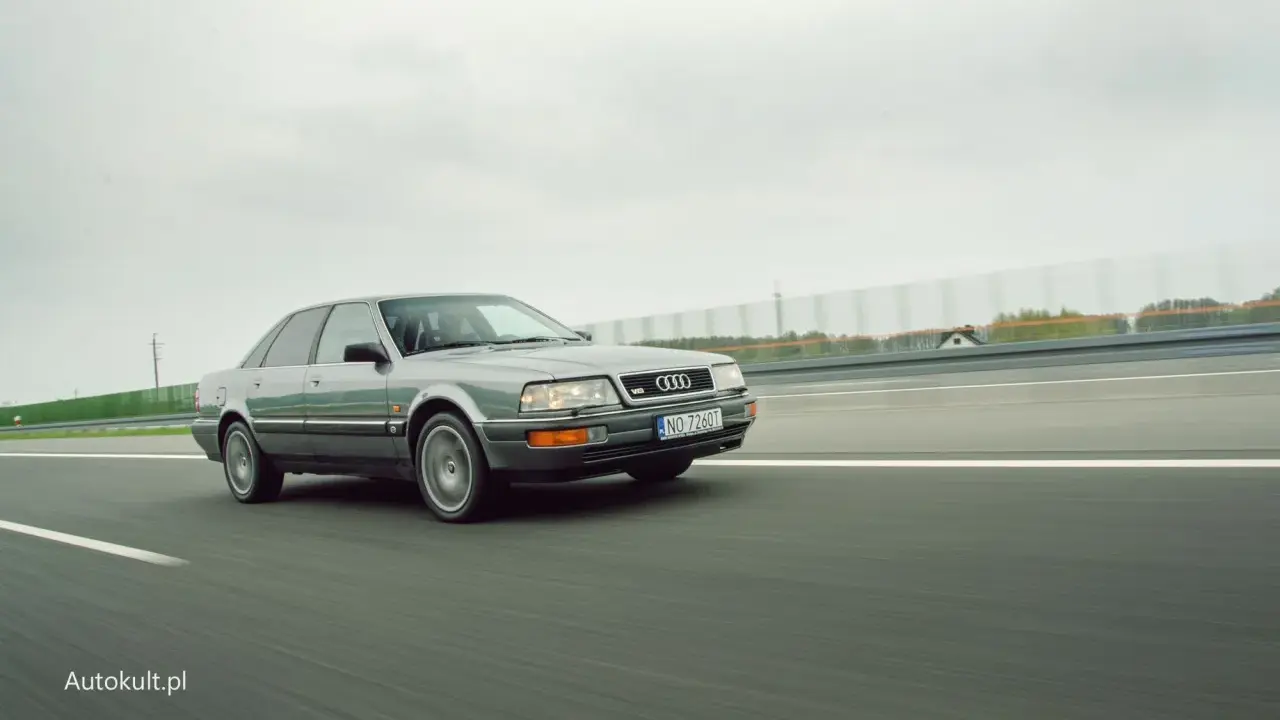 Szary Audi V8 pędzi po autostradzie, chmury na niebie i zielone drzewa w tle.