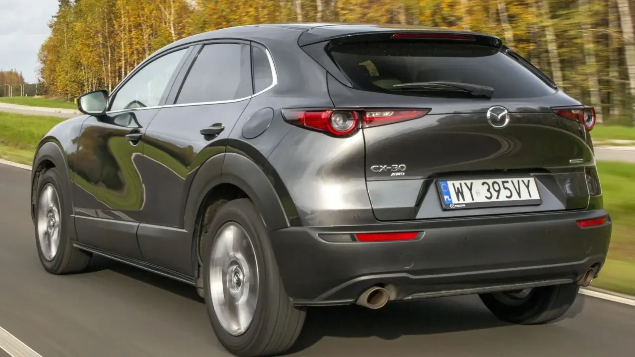 Ciemnoszara Mazda CX-30 jedzie po drodze, otoczona jesiennymi drzewami. Wady tego modelu mogą dotyczyć np. widoczności.