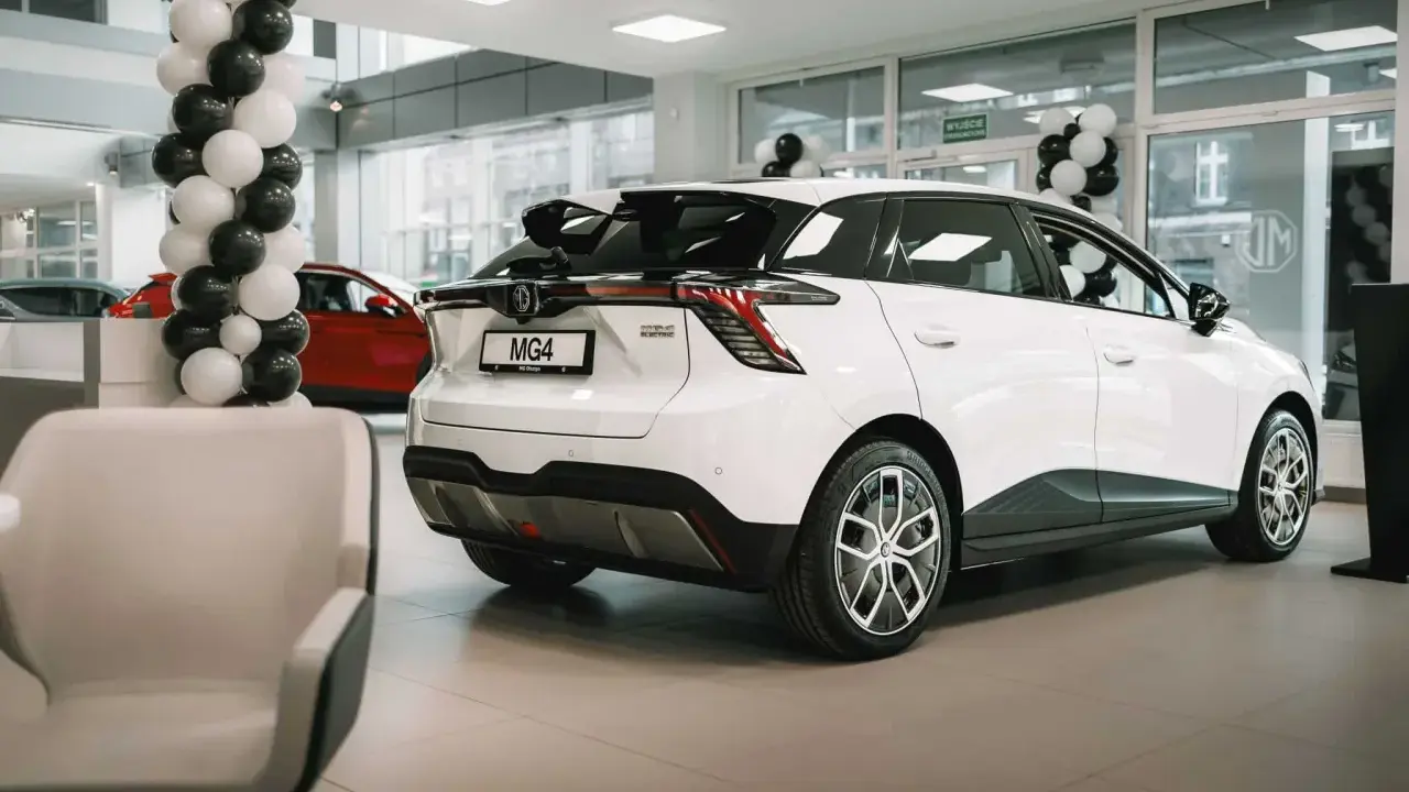 Białe auto MG 4 Electric w salonie, ozdobionym balonami. Obok czerwony samochód i wygodne fotele.