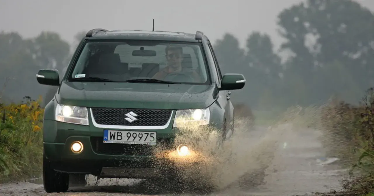Zielony Suzuki Grand Vitara 1.9 DDIS pokonuje kałużę na błotnistej drodze.