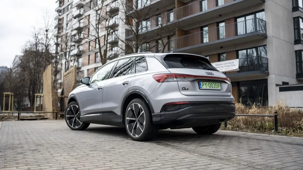 Srebrny Audi Q4 e-tron zaparkowany na brukowanym podjeździe przed nowoczesnym budynkiem mieszkalnym.