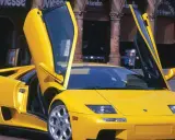 Żółty Lamborghini Diablo z otwartymi drzwiami, zaparkowany na brukowanej ulicy przed budynkami z arkadami.