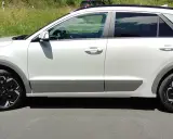 Biały Kia Niro EV z czarnymi felgami stoi na drodze, otoczony zielenią.
