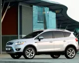 Biały Ford Kuga 2010 zaparkowany przed nowoczesnym budynkiem z przeszkloną fasadą.