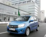 Niebieski Suzuki Celerio jedzie ulicą w mieście, mijając nowoczesne budynki i innych uczestników ruchu.