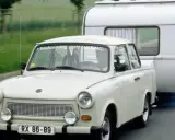 Biały Trabant ciągnie przyczepę kempingową po drodze.