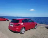 Czerwony fiat tipo sedan z napisem "SprawdzAuto24.pl" stoi nad błękitnym jeziorem w słoneczny dzień.