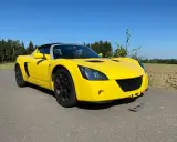 Żółty Opel Speedster na tle lasu i pól. Słońce pięknie podkreśla jego sportową sylwetkę.