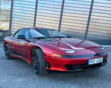 Czerwony Mitsubishi 3000GT z czarnymi felgami i przyciemnianymi szybami, zaparkowany przed nowoczesnym budynkiem.