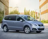 Srebrny Seat Alhambra, rodzinny van, parkuje przed nowoczesnym budynkiem z brązową elewacją.
