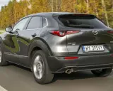 Ciemnoszara Mazda CX-30 jedzie po drodze, otoczona jesiennymi drzewami. Wady tego modelu mogą dotyczyć np. widoczności.