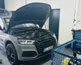 Szary Audi Q7 3.0 TDI z otwartą maską i bagażnikiem w warsztacie. Obok stoi wózek z narzędziami i silnik.