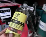Butelka z dodatkiem do oleju napędowego, z etykietą "DIESEL skydd". W tle dystrybutor paliw i pistolet do tankowania.
