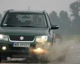 Zielony Suzuki Grand Vitara 1.9 DDIS pokonuje kałużę na błotnistej drodze.