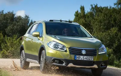 Zielony Suzuki SX4 S-Cross pokonuje teren, wzbijając kurz. Mimo że auto radzi sobie na bezdrożach, warto rozważyć jego wady przed zakupem.