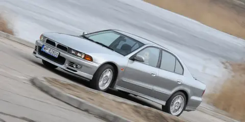 Srebrny Mitsubishi Galant z gracją pokonuje zakręt. Kierowca w okularach pewnie prowadzi auto.