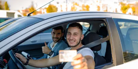 Szczęśliwy młody kierowca pokazuje swoje nowe prawo jazdy, a jego instruktor daje kciuk w górę. To jego pierwsze prawo jazdy, zdobyte w wieku 17 lat.