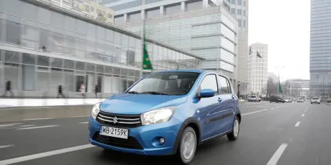 Niebieski Suzuki Celerio jedzie ulicą w mieście, mijając nowoczesne budynki i innych uczestników ruchu.