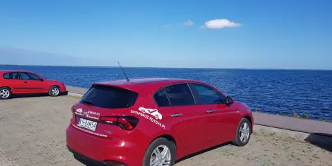 Czerwony fiat tipo sedan z napisem "SprawdzAuto24.pl" stoi nad błękitnym jeziorem w słoneczny dzień.