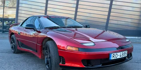 Czerwony Mitsubishi 3000GT z czarnymi felgami i przyciemnianymi szybami, zaparkowany przed nowoczesnym budynkiem.