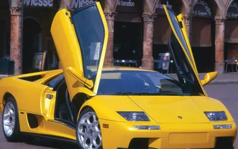 Żółty Lamborghini Diablo z otwartymi drzwiami, zaparkowany na brukowanej ulicy przed budynkami z arkadami.