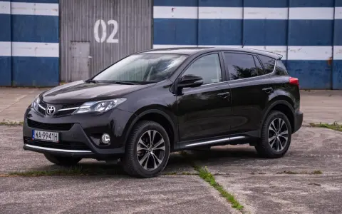 Czarny, używany Toyota RAV4 z salonu, zaparkowany przed budynkiem z niebiesko-białymi pasami.