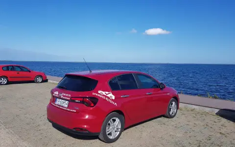 Czerwony fiat tipo sedan z napisem "SprawdzAuto24.pl" stoi nad błękitnym jeziorem w słoneczny dzień.