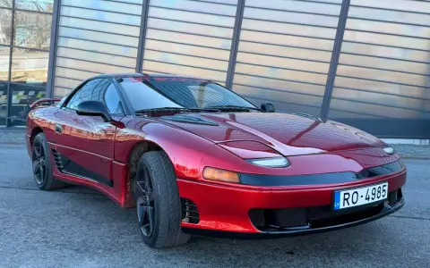 Czerwony Mitsubishi 3000GT z czarnymi felgami i przyciemnianymi szybami, zaparkowany przed nowoczesnym budynkiem.