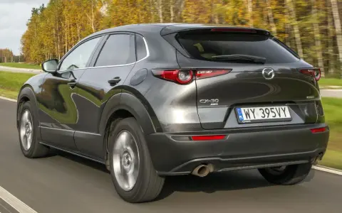 Ciemnoszara Mazda CX-30 jedzie po drodze, otoczona jesiennymi drzewami. Wady tego modelu mogą dotyczyć np. widoczności.