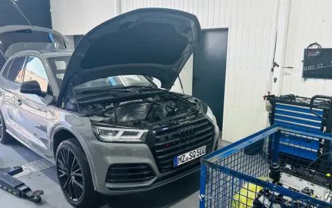 Szary Audi Q7 3.0 TDI z otwartą maską i bagażnikiem w warsztacie. Obok stoi wózek z narzędziami i silnik.