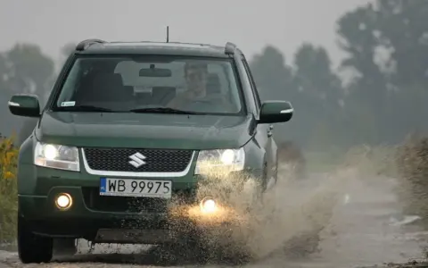 Zielony Suzuki Grand Vitara 1.9 DDIS pokonuje kałużę na błotnistej drodze.