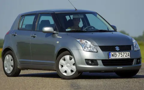 Szary Suzuki Swift 2007 z kierowcą za kierownicą, zaparkowany na drodze.