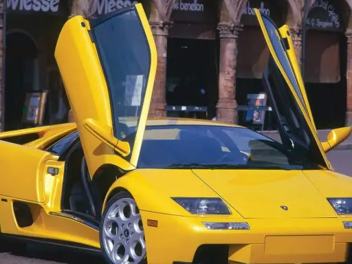Żółty Lamborghini Diablo z otwartymi drzwiami, zaparkowany na brukowanej ulicy przed budynkami z arkadami.