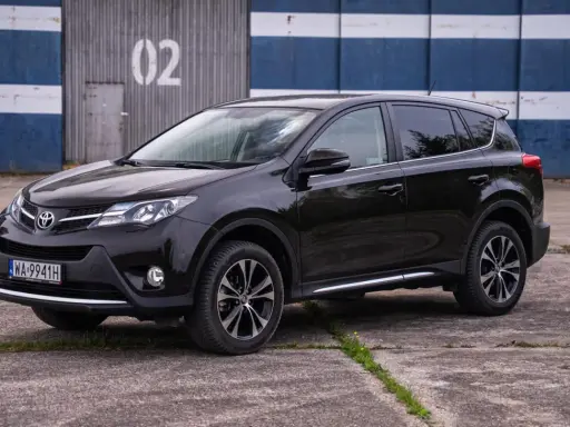 Czarny, używany Toyota RAV4 z salonu, zaparkowany przed budynkiem z niebiesko-białymi pasami.
