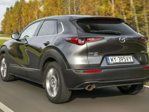 Ciemnoszara Mazda CX-30 jedzie po drodze, otoczona jesiennymi drzewami. Wady tego modelu mogą dotyczyć np. widoczności.