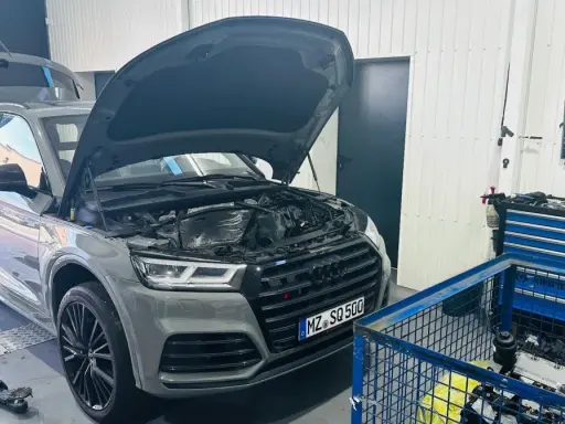 Szary Audi Q7 3.0 TDI z otwartą maską i bagażnikiem w warsztacie. Obok stoi wózek z narzędziami i silnik.