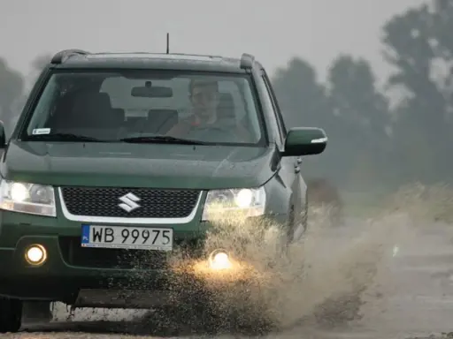Zielony Suzuki Grand Vitara 1.9 DDIS pokonuje kałużę na błotnistej drodze.