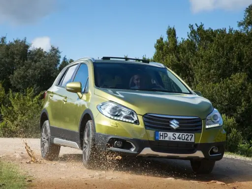 Zielony Suzuki SX4 S-Cross pokonuje teren, wzbijając kurz. Mimo że auto radzi sobie na bezdrożach, warto rozważyć jego wady przed zakupem.