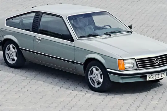 Klasyczny Opel Monza w kolorze srebrno-zielonym, z charakterystycznymi felgami i linią nadwozia, prezentuje się na tle betonowej nawierzchni.