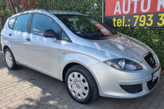Srebrny Seat Altea 1.9 TDI zaparkowany na kostce brukowej, w tle zielony żywopłot i baner z napisem "AUTO KUP".