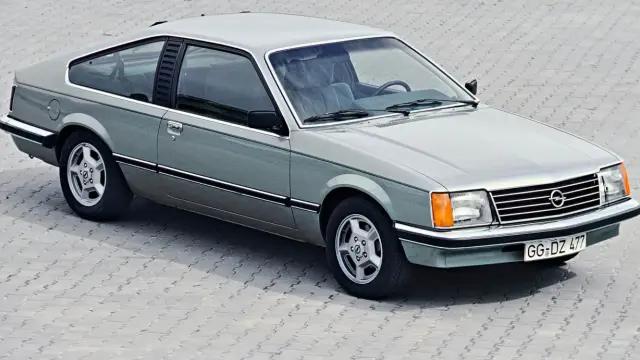 Klasyczny Opel Monza w kolorze srebrno-zielonym, z charakterystycznymi felgami i linią nadwozia, prezentuje się na tle betonowej nawierzchni.